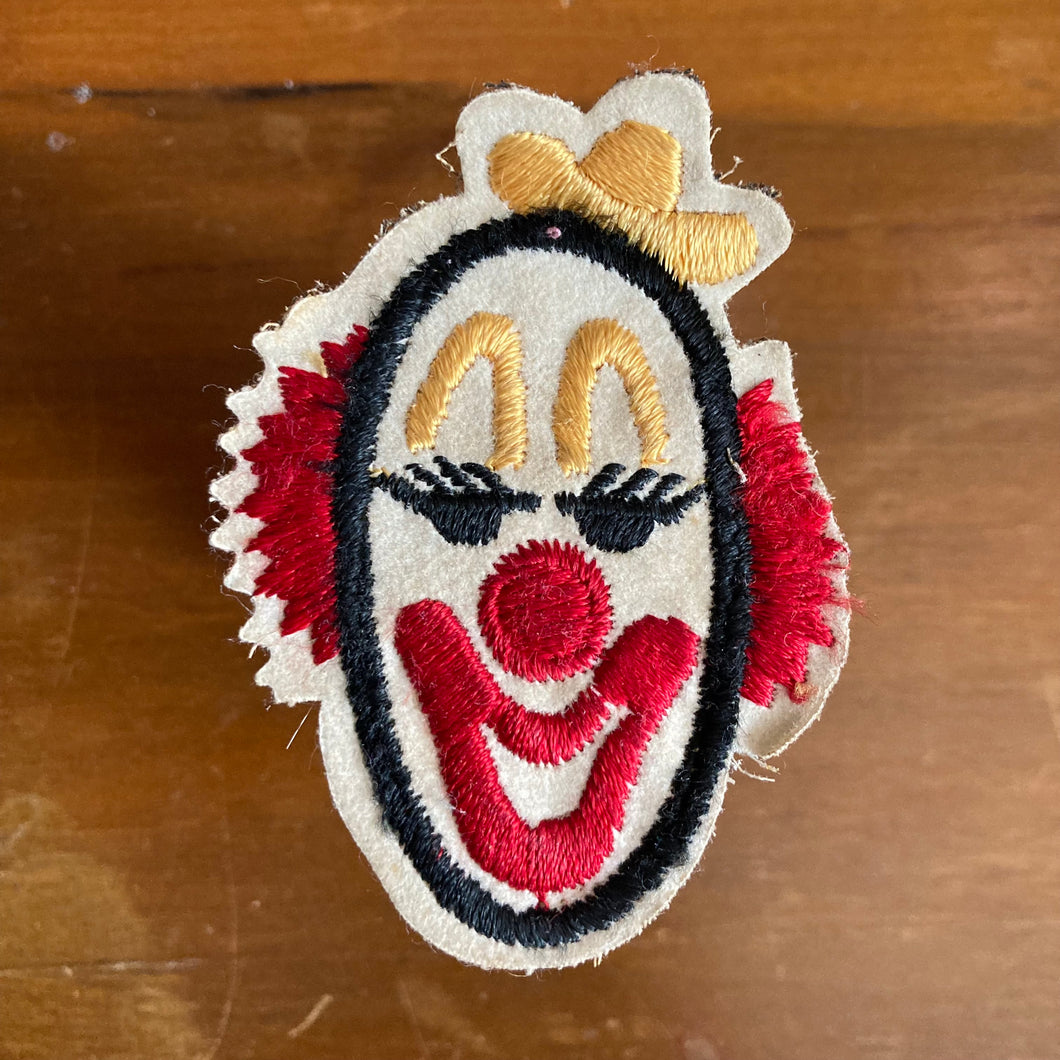 Vintage 1970's McDonalds patch with a retro clown head logo. An embroidered clown face patch on a beige fabric background, featuring red ruffles, a red nose and smile, black outlines, and yellow bow details, placed on a wooden surface.